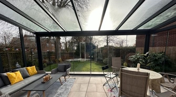 Sunlit interior of a glass enclosure with garden view, sofa, and dining table