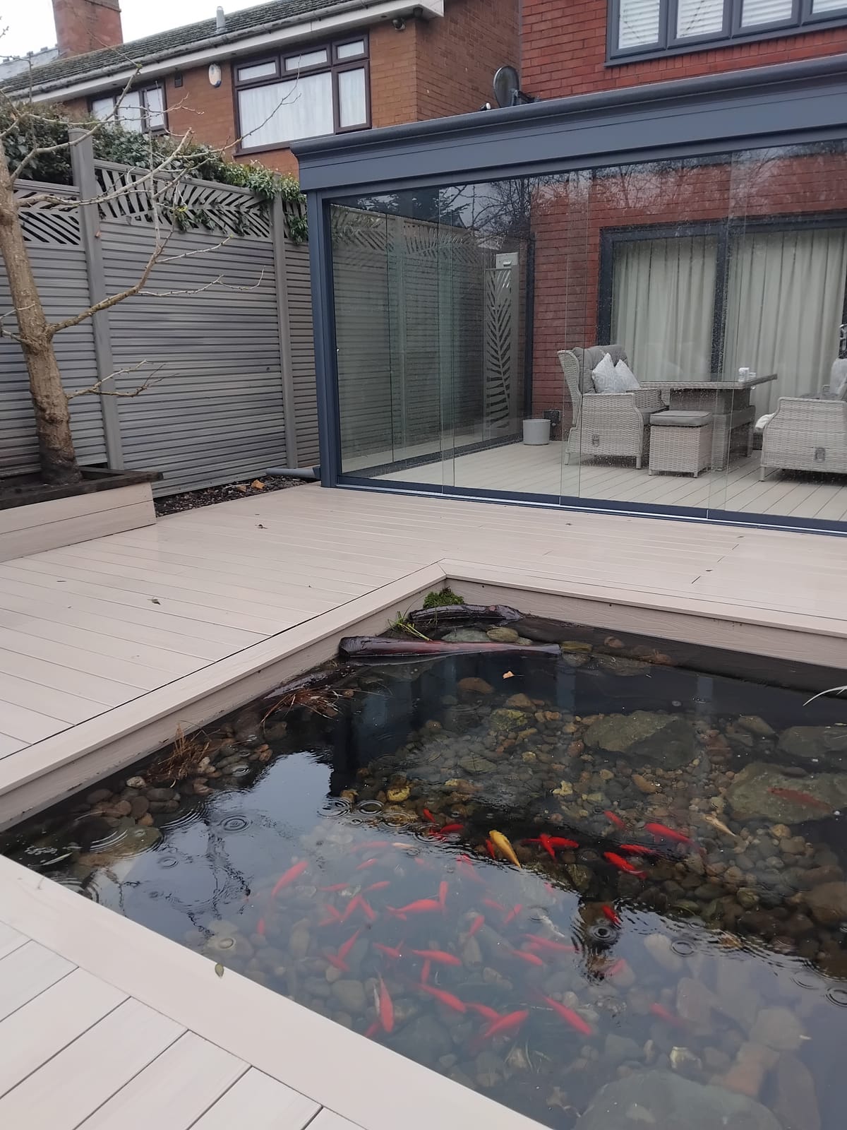 Glass garden room with frameless sliding glass walls beside a koi pond, attached to a red-brick house